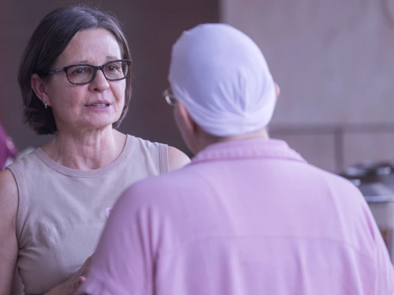 Two people talk outdoors. One, facing camera, has short brown straight hair and black rectangle classes. The other is wearing a scarf on her head.