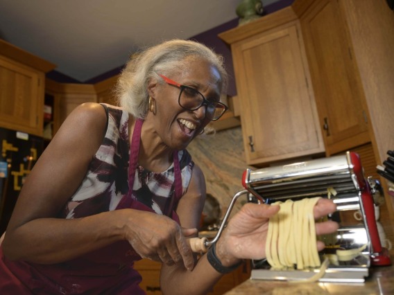 A person pulls linguine from a pasta-making machine. She has short gray hair and red glasses and is laughing.