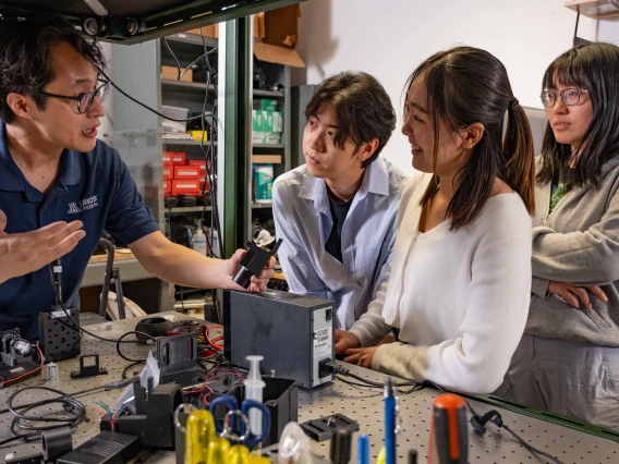 A professor with dark, straight short hair and glasses shows a small black device to three students who are watching the speaker intently.  