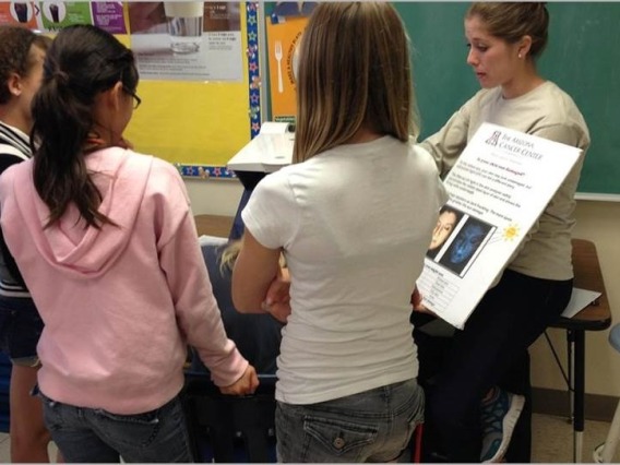 Four students encircle a person presenting a chart on sun safety. The speaker has her hair pulled up and is wearing a white long sleeve sweater.
