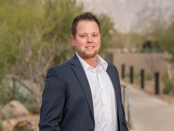 Person stands outside the University of Arizona Cancer Center. he is wearing a gray jacket and white button-down shirt and has short dark hair and a beard. He is smiling.