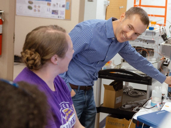 Two people in a lab look at each other and smile. One is standing with a blue button down shirt and short hair, and one person has long brown hair in a clip and is wearing a purple T-shirt