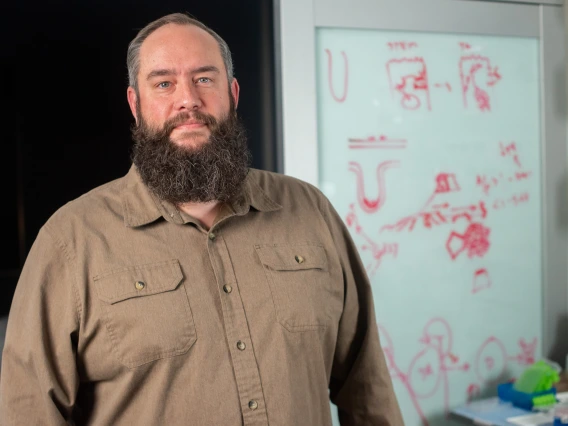 Person smiles into the camera. He has a dark beard and is wearing a brown button-down shirt.