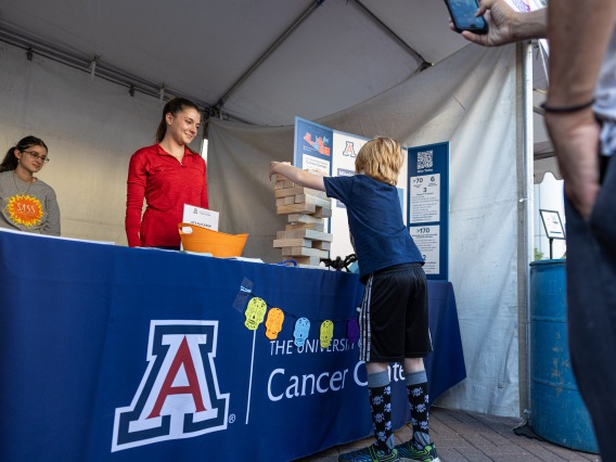 Two students assist at the Skin Cancer event booth. A young boy plays a giant Jenga on the table while his father looks on and take a photo.