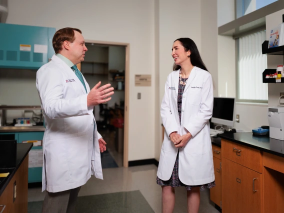 Two researchers in white coats talk in their lab.