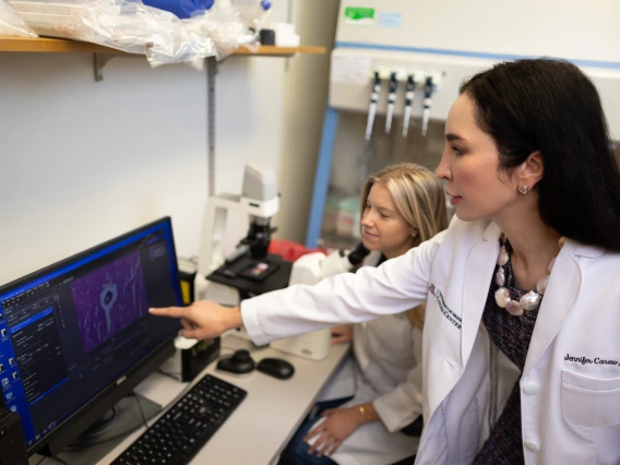 A person in a lab coat points to a computer screen as a student looks at the screen.