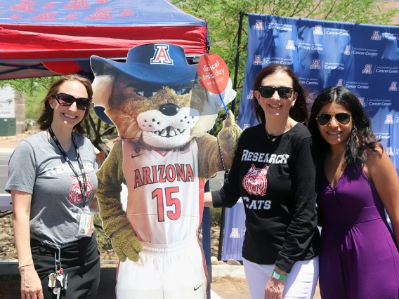 Three people pose with a cutout of the U of A mascot, Wilbur Wildcat outdoors in front of the North Cancer Center