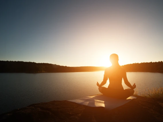 Silhouette of a person sitting cross legged on yoga mat in prayer position in front of a lake at sunset.