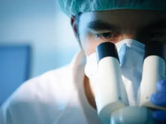 Researcher looks into a microscope wearing mask, hairnet and lab coat.