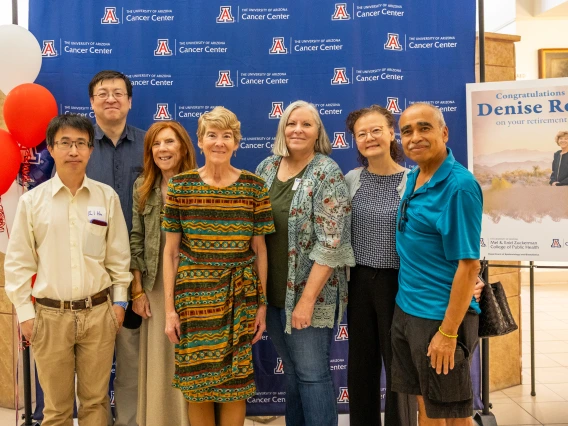 Denise Roe poses for a photo with six of her colleagues in front of a U of A Cancer Center blue backdrop next to a sign announcing her retirement.