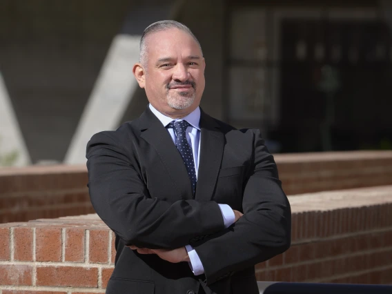 An outdoor portrait of John Ruiz with arms crossed wearing a suit and tie.