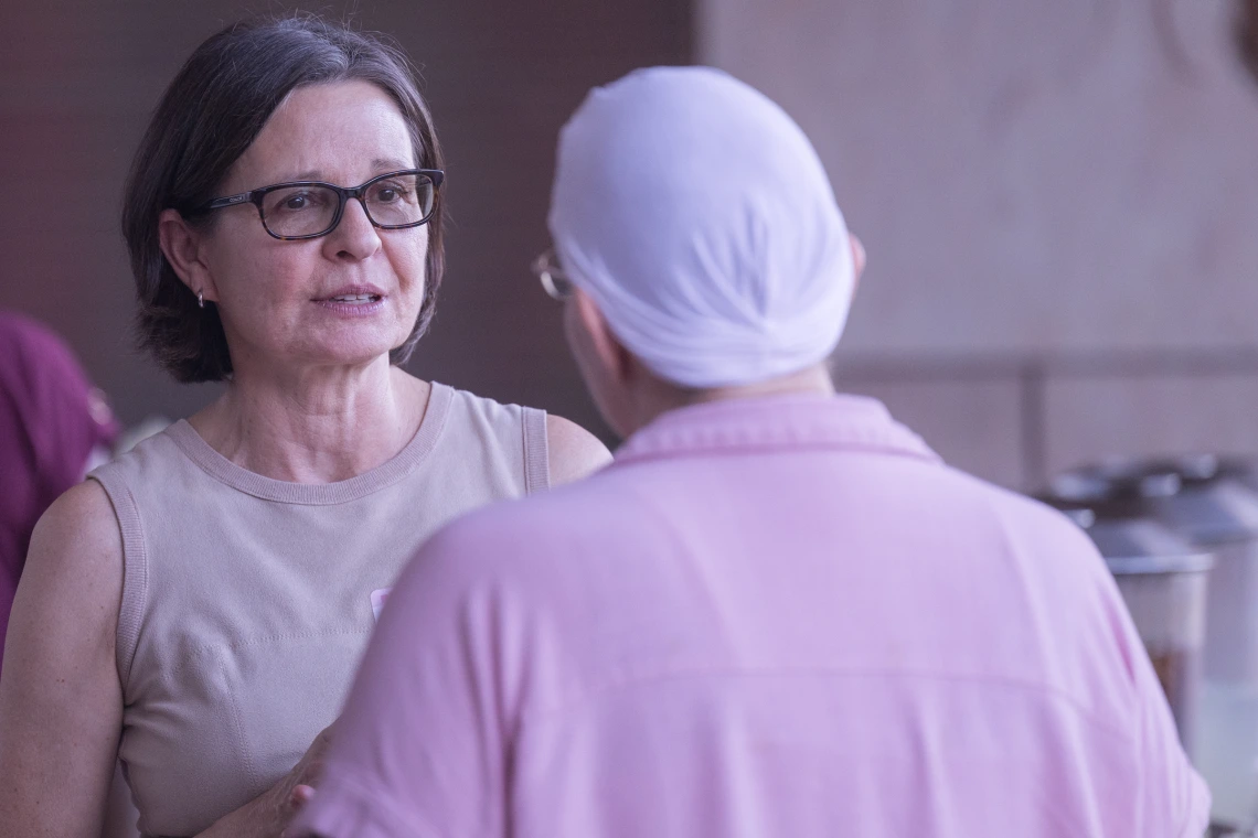 Two people talk outdoors. One, facing camera, has short brown straight hair and black rectangle classes. The other is wearing a scarf on her head.