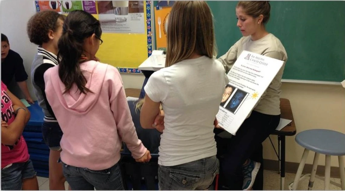 Four students encircle a person presenting a chart on sun safety. The speaker has her hair pulled up and is wearing a white long sleeve sweater.