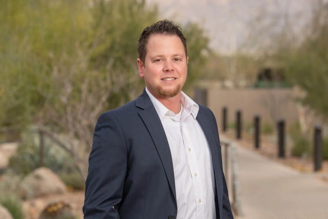 Person stands outside the University of Arizona Cancer Center. he is wearing a gray jacket and white button-down shirt and has short dark hair and a beard. He is smiling.