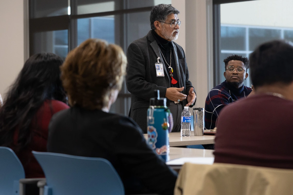 Person standing talks to a group sitting at a rectangular table.