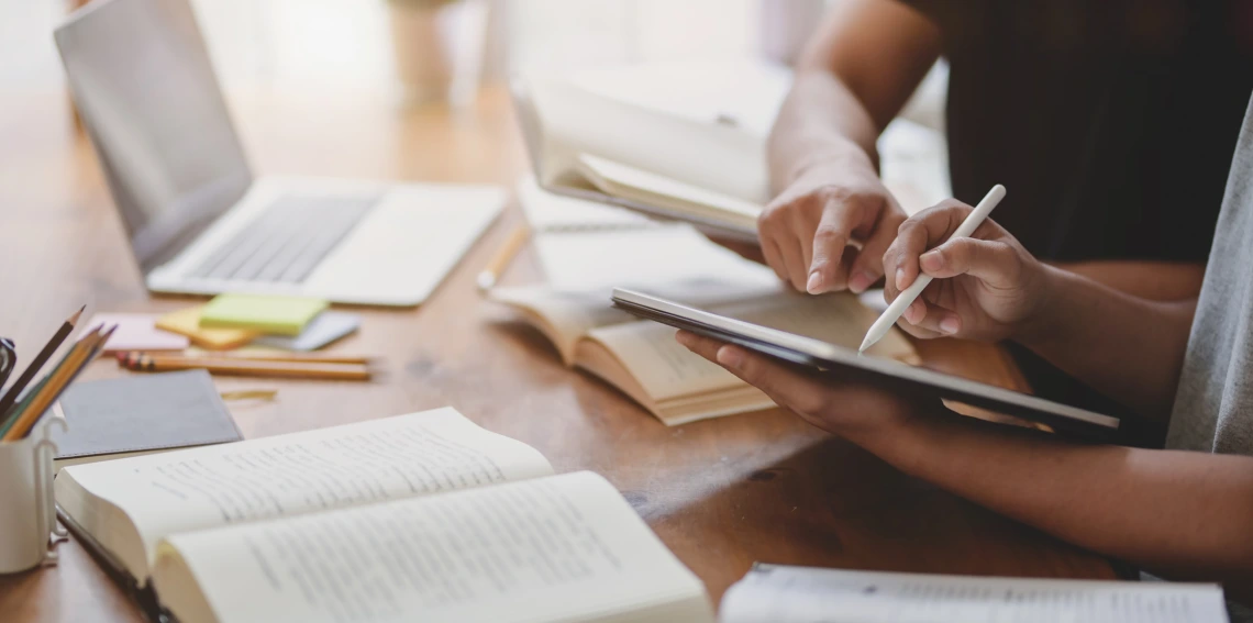 two people sit at a table next to each other. One is pointing to an iPad while the other works on a document with an Apple Pencil 