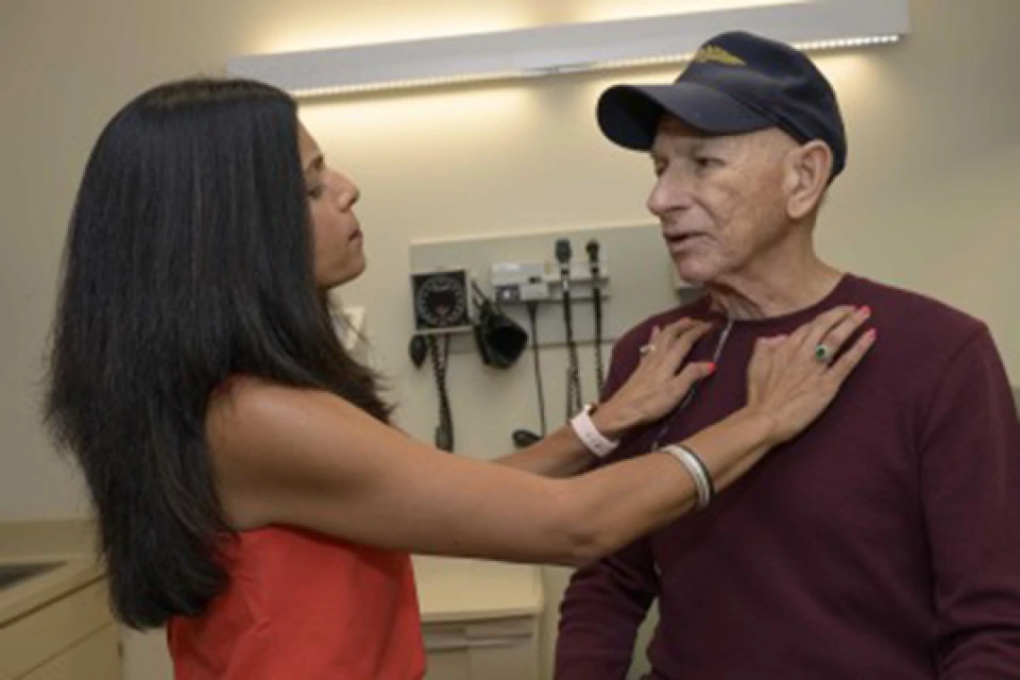 A doctor examines a patient in an examining room. The patient is wearing a Navy ball cap.