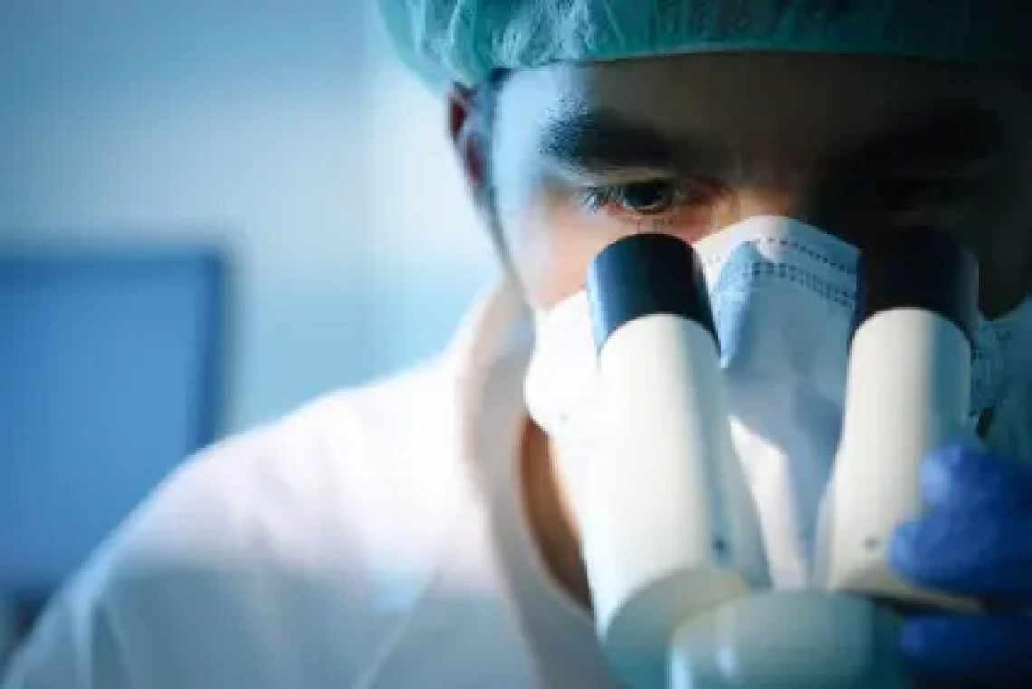 Researcher looks into a microscope wearing mask, hairnet and lab coat.