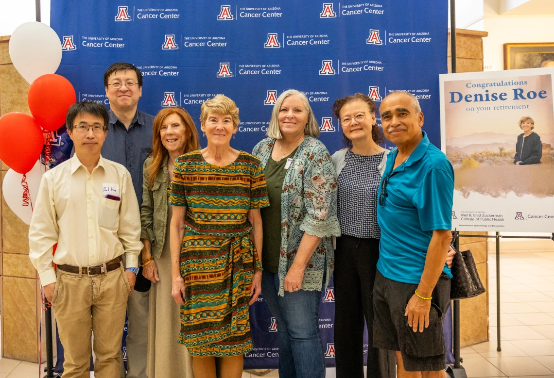 Denise Roe poses for a photo with six of her colleagues in front of a U of A Cancer Center blue backdrop next to a sign announcing her retirement.