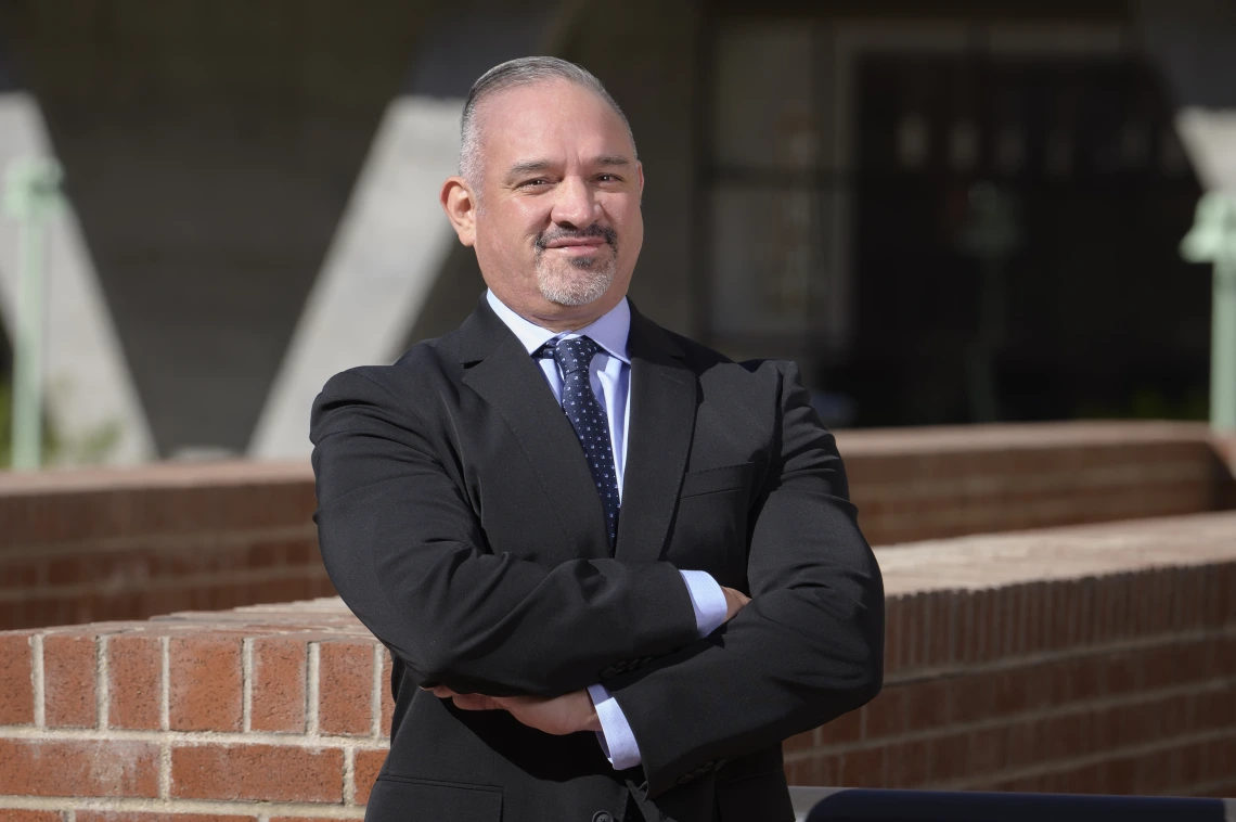 An outdoor portrait of John Ruiz with arms crossed wearing a suit and tie.