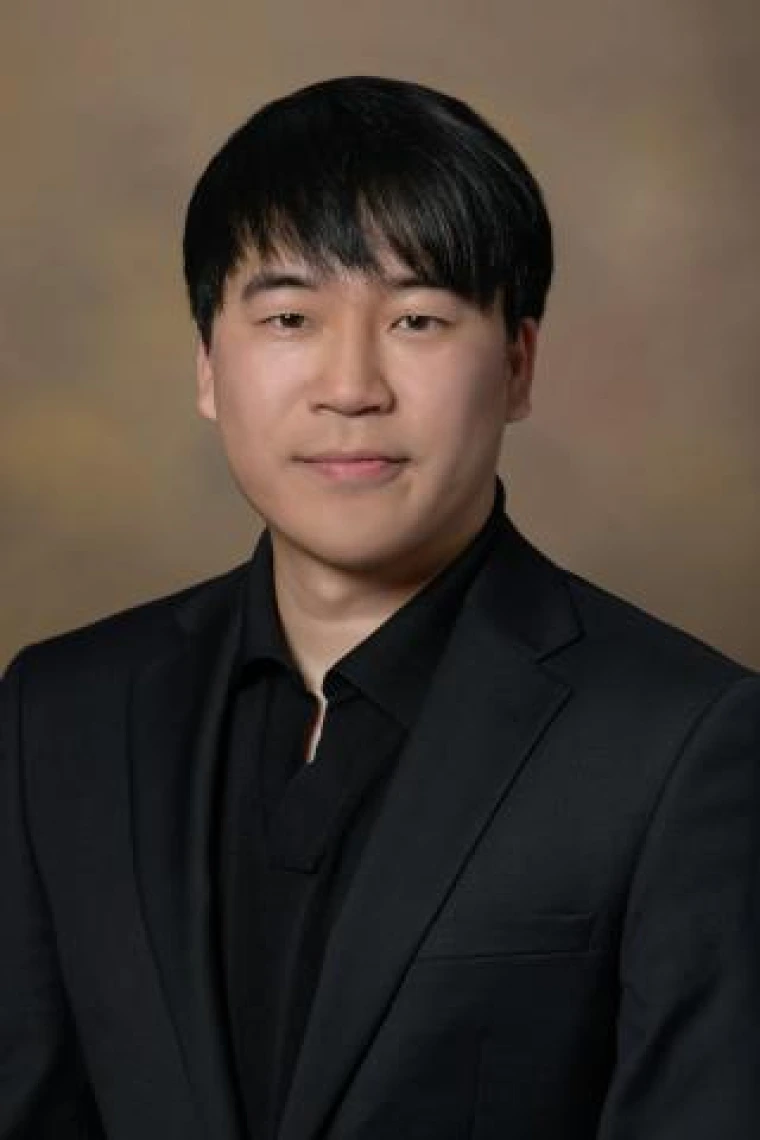 Portrait of person with dark, short straight hair. He is wearing a black button-down shirt and black jacket.