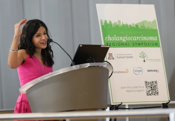 Person speaks at a podium. She has dark, wavy shoulder-length hair and has her arm lifted up to indicate height.