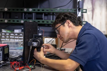 Person works on a prototype devices. He has short dark hair and glasses and is wearing a blue short-sleeved polo shirt.