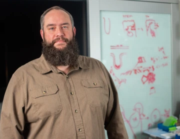 Person smiles into the camera. He has a dark beard and is wearing a brown button-down shirt.