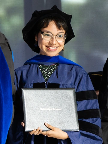 Person wearing a graduation cap and gown poses with their diploma.