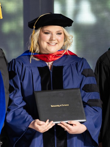 Person wearing a graduation cap and gown poses with their diploma