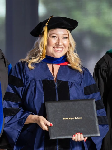 Person poses with their diploma. They are wearing a graduation cap and gown and are smiling.