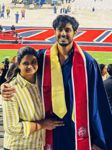 A graduate student poses for a photo in his graduation gown and stoles with his mom. He is in the stands above the University of Arizona football field.