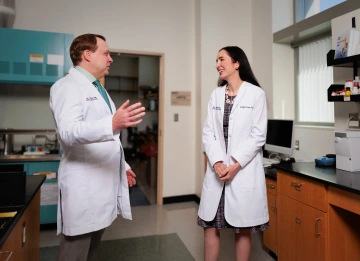 Two researchers in white coats talk in their lab.