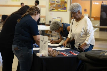 Two community members sign in during a Let's Taco 'Bout Cancer event while two community health workers assist them behind a table.