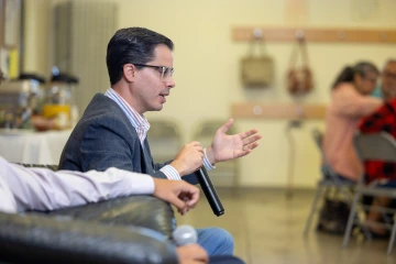 Dr. Alejandro Recio Boiles holds a microphone while seated in an armchair inside a community center.