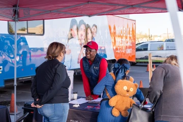 Namoonga Mantina stands behind a table next to the mobile mammography unit assisting a participant.