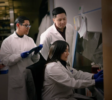 A person works inside a hood in a lab while two people in lab coast look on.