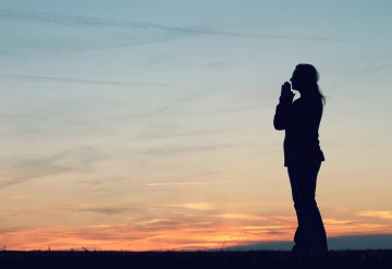 Silhouette of person facing sunset with hands clasped.