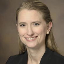 A person smiles with blond hair pulled back. She is wearing gold button earrings and a black jacket.