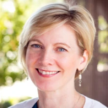 A person smiles with short blond hair and long hanging earrings. She is wearing a white blouse.