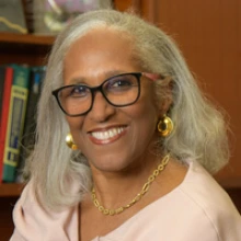 A person is smiling with shoulder-length gray hair. She is wearing black glasses and gold hoop earrings and a pink blouse.