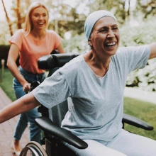 Person in wheelchair wearing a head scarf with arms extended pushed by their daughter 