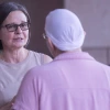 Two people talk outdoors. One, facing camera, has short brown straight hair and black rectangle classes. The other is wearing a scarf on her head.