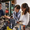 A professor with dark, straight short hair and glasses shows a small black device to three students who are watching the speaker intently.  