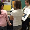 Four students encircle a person presenting a chart on sun safety. The speaker has her hair pulled up and is wearing a white long sleeve sweater.