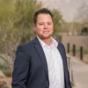 Person stands outside the University of Arizona Cancer Center. he is wearing a gray jacket and white button-down shirt and has short dark hair and a beard. He is smiling.