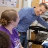 Two people in a lab look at each other and smile. One is standing with a blue button down shirt and short hair, and one person has long brown hair in a clip and is wearing a purple T-shirt