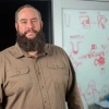 Person smiles into the camera. He has a dark beard and is wearing a brown button-down shirt.