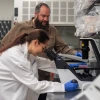 A person in a lab coat with long brown hair in a ponytail  and blue latex gloves looks into a microscope. Another person with a beard and brown button down shirt watches from the side.