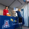 Two students assist at the Skin Cancer event booth. A young boy plays a giant Jenga on the table while his father looks on and take a photo.